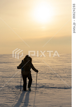 Human silhouette in hooded down jacket and ski poles walks througth snow-covered plain at sunset, vertical photo, selected focus. 86624288