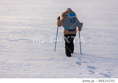 Human in beige down jacket with blue backpack and ski poles walking in the snow-covered plain leave traces, back view, selected focus. 86624291