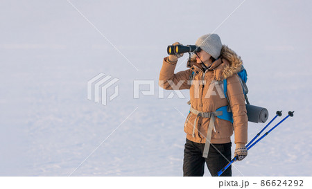 Woman in down jacket with a backpack and ski poles in hand looks through binoculars standing on a snowy plain in winter, selected focus. 86624292