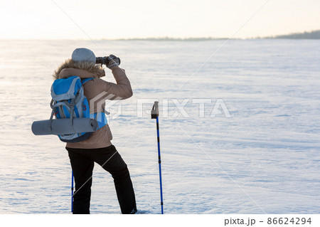 Human in beige down jacket with a blue backpack and ski poles looks through binoculars in the middle of a snowy plain in winter, back view, selected focus. 86624294