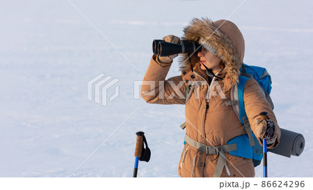 Woman in yellow hooded down jacket with a backpack and ski poles looks through binoculars standing on a snowy plain in winter, waist up, selected focus. 86624296
