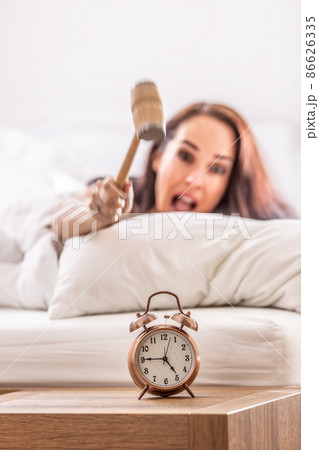 Smashing of alarm clock with the hammer by a woman reaching out from the bed Smashing of alarm clock with the hammer by a woman reaching out from the bed 86626335