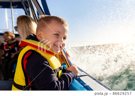 Portrait of cite little blond happy excited smiling caucasian boy wear lifevest enjoy sailing on motor boat sea against blue sky and water splash wave sun backlit. Summer travel vacation recreation 86629257