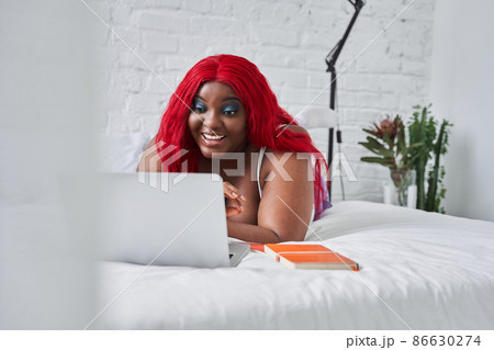 Red haired woman laughing while laying on the bed and working at the laptop Red haired woman laughing while laying on the bed and working at the laptop 86630274