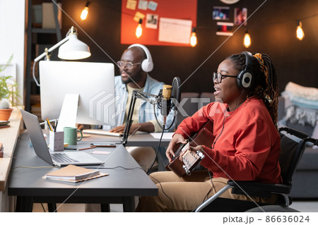 African woman with disability sitting on wheelchair playing guitar and singing a song to the microphone with man using computer in the background African woman with disability sitting on wheelchair playing guitar and singing a song to the microphone with man using computer in the background 86636024