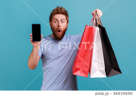 Portrait of astonished surprised bearded man standing with open mouth showing shopping bags and smart phone with blank screen for advertisement. Indoor studio shot isolated on blue background. Portrait of astonished surprised bearded man standing with open mouth showing shopping bags and smart phone with blank screen for advertisement. Indoor studio shot isolated on blue background. 86639690