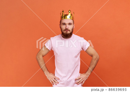 Portrait of bearded man egoistically looking at camera, posing with crown on head, pretending to be king, privileged status, wearing pink T-shirt. Indoor studio shot isolated on orange background. 86639693