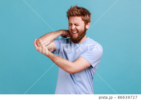 Portrait of bearded man massaging aching elbow, frowning from acute pain, strain and strain problem, trapped nerves, rheumatoid arthritis. Indoor studio shot isolated on blue background. 86639727