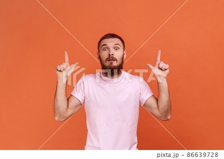 Wow, look, crazy advertisement. Portrait of bearded man pointing up at empty place for ad content and expressing astonishment, wearing pink T-shirt. Indoor studio shot isolated on orange background. 86639728
