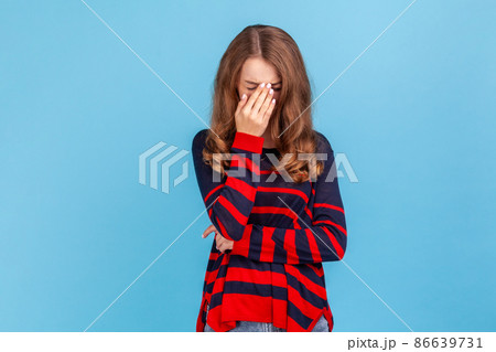 Depressed upset young woman wearing striped casual style sweater bowing her head and crying, experiencing desperate sorrowful feelings. Indoor studio shot isolated on blue background. Depressed upset young woman wearing striped casual style sweater bowing her head and crying, experiencing desperate sorrowful feelings. Indoor studio shot isolated on blue background. 86639731