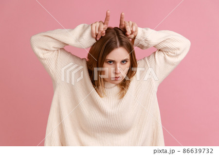 Portrait of aggressive blond woman making bull horns gesture over her head and frowning face, expressing anger threat, wearing white sweater. Indoor studio shot isolated on pink background. Portrait of aggressive blond woman making bull horns gesture over her head and frowning face, expressing anger threat, wearing white sweater. Indoor studio shot isolated on pink background. 86639732