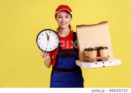 Portrait of smiling courier woman holding wall clock and coffee with pizza box, express delivery in time, wearing overalls and red cap. Indoor studio shot isolated on yellow background. Portrait of smiling courier woman holding wall clock and coffee with pizza box, express delivery in time, wearing overalls and red cap. Indoor studio shot isolated on yellow background. 86639733