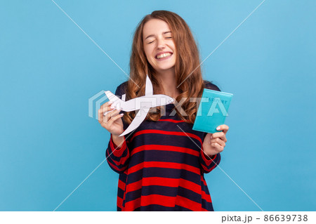 Extremely satisfied woman wearing striped casual style sweater, holding passport and paper plane in hands, rejoicing the vacation abroad, adventures. Indoor studio shot isolated on blue background. 86639738