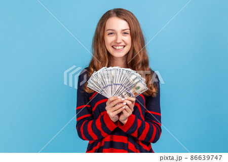 Satisfied woman wearing striped casual style sweater, holding fan of dollar bills and looking ar camera with toothy smile, lottery winner. Indoor studio shot isolated on blue background. 86639747