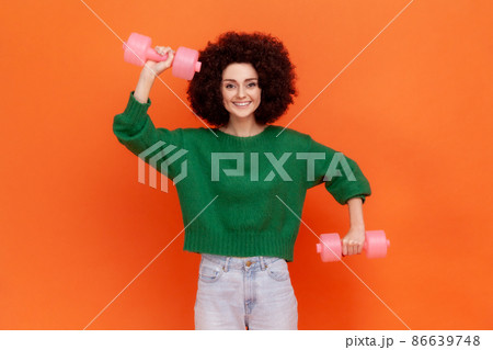 Happy positive sporty woman with Afro hairstyle wearing green casual style sweater doing exercises for biceps, using dumbbells, health care. Indoor studio shot isolated on orange background. 86639748
