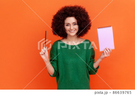 Portrait of happy woman with Afro hairstyle wearing green casual style sweater standing holding paper notebook and pen, looking at camera with smile. Indoor studio shot isolated on orange background. Portrait of happy woman with Afro hairstyle wearing green casual style sweater standing holding paper notebook and pen, looking at camera with smile. Indoor studio shot isolated on orange background. 86639765