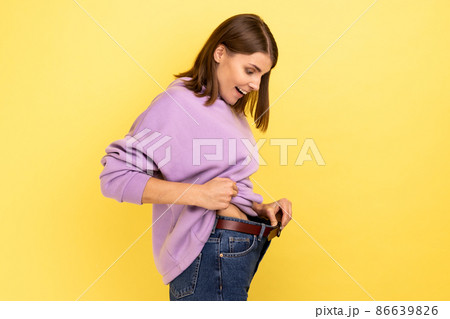 Side view of dark haired happy woman showing successful weight loss, expressing happiness, diet concept, wearing purple hoodie. Indoor studio shot isolated on yellow background. Side view of dark haired happy woman showing successful weight loss, expressing happiness, diet concept, wearing purple hoodie. Indoor studio shot isolated on yellow background. 86639826