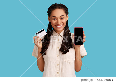 Smiling woman with black dreadlocks showing mobile phone, holding credit bank card, online banking, looking at camera, wearing white shirt. Indoor studio shot isolated on blue background. 86639863