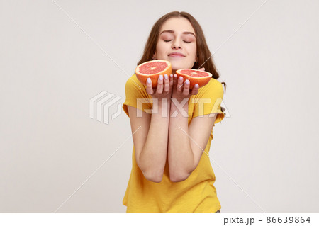 Portrait of beautiful teenager girl with brown hair smelling half slice grapefruit, enjoying tasty fresh juicy vitamin fruits, healthy lifestyle. Indoor studio shot isolated on gray background. 86639864