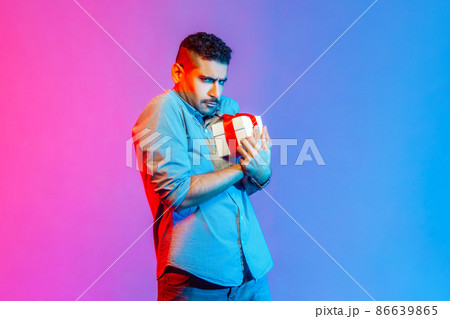 Portrait of man in shirt holding present box in hands, being greedy to share gifts, looking at camera with frowning face. Indoor studio shot isolated on colorful neon light background. 86639865
