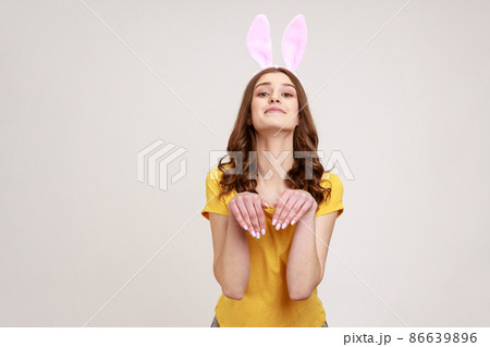 Portrait of joyful beautiful woman of young age in yellow T-shirt behaving childish humorous with bunny ears and hands, entertaining and fooling. Indoor studio shot isolated on gray background. Portrait of joyful beautiful woman of young age in yellow T-shirt behaving childish humorous with bunny ears and hands, entertaining and fooling. Indoor studio shot isolated on gray background. 86639896