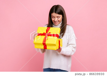 Positive happy dark haired girl looking inside wrapped present box, open gift, expressing positive, wearing white casual style sweater. Indoor studio shot isolated on pink background. Positive happy dark haired girl looking inside wrapped present box, open gift, expressing positive, wearing white casual style sweater. Indoor studio shot isolated on pink background. 86639930