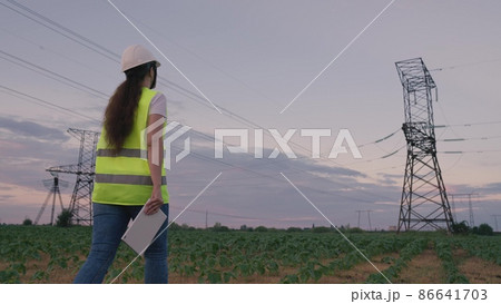 high energized towers against backdrop of sunset in the sky. woman architect hard hat with tablet designing place to control equipment. young woman energy voltage specialist. electric towers at sunset 86641703