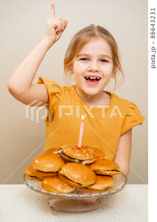 a funny kid little girl makes a wish and blows out a candle on a hamburger cake. a funny kid little girl makes a wish and blows out a candle on a hamburger cake. 86643231