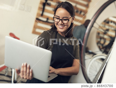 Emails are how I stay in touch with clients. Shot of an attractive young woman standing alone in her bicycle shop and using her laptop. 86643700