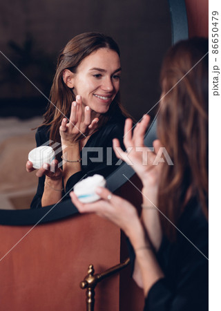 Young woman in front of the mirror putting cream on her face Young woman in front of the mirror putting cream on her face 86650479