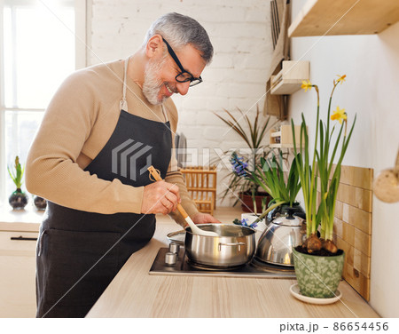 Happy senior man in apron cooking in kitchen at home, standing near stove and stirring sauce in pan Happy senior man in apron cooking in kitchen at home, standing near stove and stirring sauce in pan 86654456