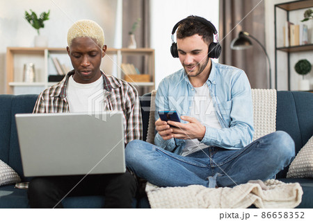 Multiracial same sex couple resting on couch with gadgets Multiracial same sex couple resting on couch with gadgets 86658352