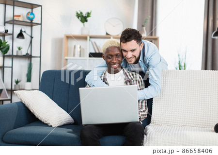 Happy same sex couple sitting on couch and using laptop Happy same sex couple sitting on couch and using laptop 86658406