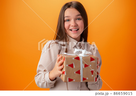 Cute teen girl holding a Christmas gift box against orange background. 86659015