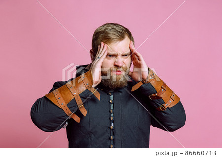 Portrait of young bearded man in image of medieval warrior, archer suffering from pain, touching temples in headache isolated over pink background 86660713