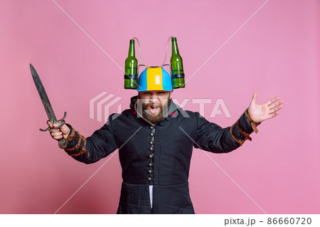 Shouting. Half-length portrait of young bearded man in image of medieval warrior, archer in beer helmet with big knife isolated over pink studio background. Beer festival concept 86660720