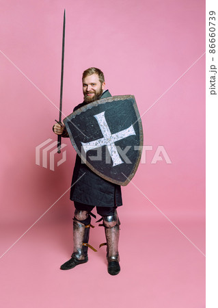 Portrait of young brave man, medieval warrior or archer with sword and protective shield isolated over pink studio background 86660739