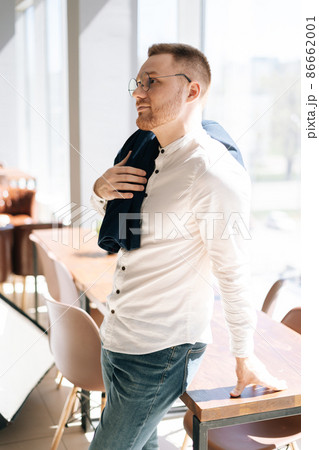 Vertical portrait of handsome confident young business man in elegance eyeglasses wearing stylish white clothes standing posing in office, by window and thinking looking away. 86662001
