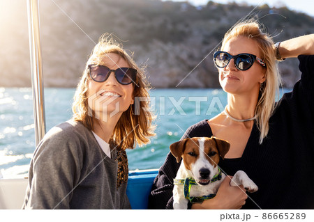 Portrait two young adult beautiful pretty caucasian woman enjoy sailing family cute adorable little jack russel dog on yacht boat against blue water bright summer day. Travel sea tourism with pets Portrait two young adult beautiful pretty caucasian woman enjoy sailing family cute adorable little jack russel dog on yacht boat against blue water bright summer day. Travel sea tourism with pets 86665289