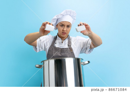 Portrait of young caucasian woman, lady-cook, chef in white uniform with saucepan isolated on blue background. Funny meme emotions Portrait of young caucasian woman, lady-cook, chef in white uniform with saucepan isolated on blue background. Funny meme emotions 86671084