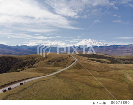 Aerial view of amazing road with view of the Caucasian ridge and Mount Elbrus. Highway runs through green meadow. Light clouds cover the white snowy peaks of Elbrus and the blue sky Aerial view of amazing road with view of the Caucasian ridge and Mount Elbrus. Highway runs through green meadow. Light clouds cover the white snowy peaks of Elbrus and the blue sky 86672890