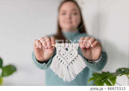 Closeup hand of girl, she shows macrame. Natural cotton threads and wooden beads. Handmade macrame belt in work process. Female hobby. Wall hanging decor. Closeup hand of girl, she shows macrame. Natural cotton threads and wooden beads. Handmade macrame belt in work process. Female hobby. Wall hanging decor. 86675315