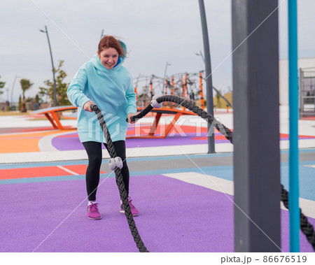 Caucasian woman in a mint sweatshirt is training with battle ropes at the sports ground.  86676519