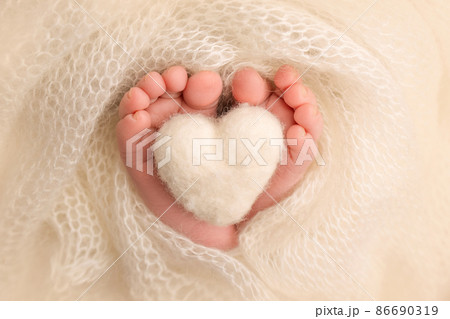The tiny foot of a newborn baby. Soft feet of a new born in a white wool blanket. Close up of toes, heels and feet of a newborn. Knitted white heart in the legs of a baby. Macro photography. The tiny foot of a newborn baby. Soft feet of a new born in a white wool blanket. Close up of toes, heels and feet of a newborn. Knitted white heart in the legs of a baby. Macro photography. 86690319