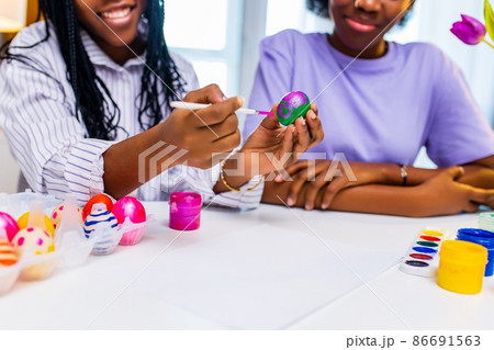 multiracial two women painting eggs with cute pink rabbit costume ears in apartment 86691563