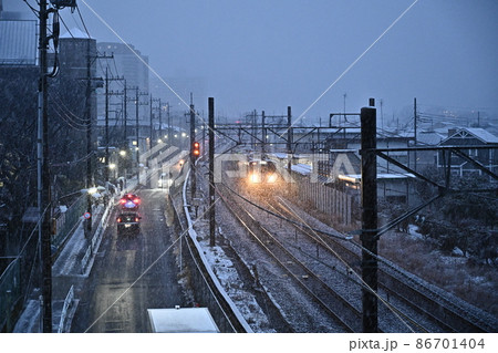 雪の夕暮れ鉄道風景　近づく電車のヘッドライト　線路横を走るトラックの屋根　薄ら白くなった街並み 86701404