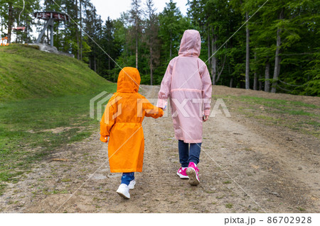 Pair of cute little caucasian sibling kids in bright waterproof raincoats walking under rain in scenic moody dirt forest road outside. Autumn weather outdoor hiking. Children friendship and support 86702928