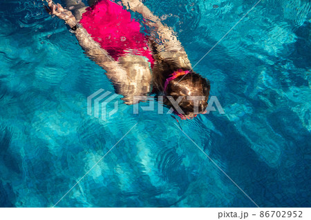Above top view of little cute kid girl in sport pink swimsuit and goggles diving underwater in clear blue water of indoor swimming pool at dark evening time. Children watersport healthy activities 86702952