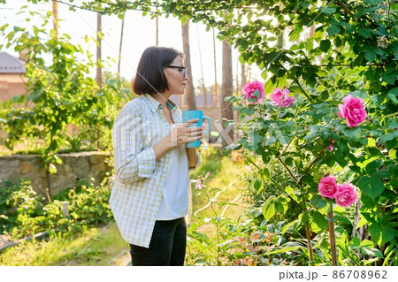 Middle-aged happy woman relaxing in garden with a cup in her hands, enjoying blooming rose Middle-aged happy woman relaxing in garden with a cup in her hands, enjoying blooming rose 86708962