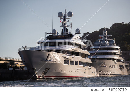 Two huge yachts are in port of Monaco at sunset, sun reflection are on glossy boards of motor boats, the chrome plated handrail, megayacht is moored in marina, mountain is on background Two huge yachts are in port of Monaco at sunset, sun reflection are on glossy boards of motor boats, the chrome plated handrail, megayacht is moored in marina, mountain is on background 86709759